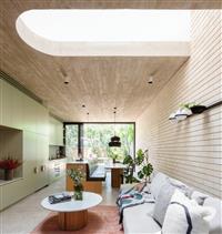 Interior kitchen and living space with large curved skylight looking down upon living area with grey couch and bench dining seating green cabinetry stretches along left wall leading to leafy outdoor area through large window at rear