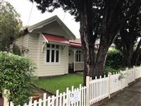 Streetfront of creme residential building with red feature roofing white framed windows white picket fence grassed area hedging and two large trees in front yard