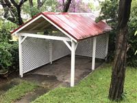 Creme outdoor carport with red tin roof white lattice surrounded by garden and one large tree and grass in foreground