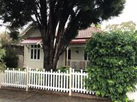 Streetfront of creme residential building taken slightly from the side with red feature roofing white framed windows white picket fence grassed area hedging and one large tree in front yard