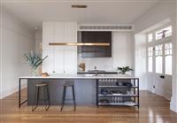 Interior kitchen space with white and black cabinetry two stools at breakfast bar island bench eucalyptus pieces in vase and plate of croissants sit on the kitchen bench and a long rectangular brass lighting fixture hangs above