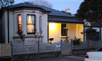 Evening streetfront exterior creme residential building with single light fixture at entry way with yellow features and grey roofing with white picket fence sandstone walling with a few small bushes dotted in shallow front yard