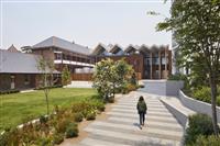 Outdoor space with person walking along grey pavered path leading from right foreground to the centre of the image where the entrance to a timber panelled building with two brick buildings on the left and green area with grass and scattered small trees and bushes