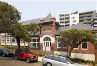 Streetfront exterior of restored red brick building featuring words Marrickville Hospital with creme features and various windows plus towering white and grey building in background and three cars in foreground