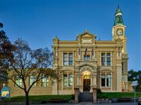 Evening scene of yellow and creme heritage building with words Town Hall at centre top of building plus three flagpoles above entry way spire like clock tower to right one large tree in left foreground