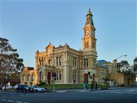 Late afternoon scene taken from light right of yellow and creme heritage building with words Town Hall at centre top of building plus three flags above entry way and spire like clock tower plus road intersection in foreground with two cars and tree people waiting