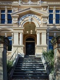 Exterior of yellow and creme building entry way with stairs coming down to centre foreground work Sydney Smith Mayor 1888 above entry way with three fingered light above