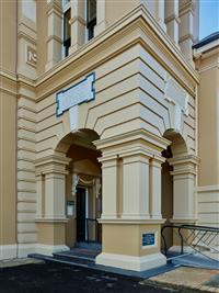 Yellow and creme building exterior showing side entry way with blue featured plaque above plus dark grey ground leading up two steps to entry way