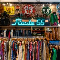 Several racks of vintage shirts, hats and boots lined up in a store, with vintage signs on the wall including a neon sign reading 