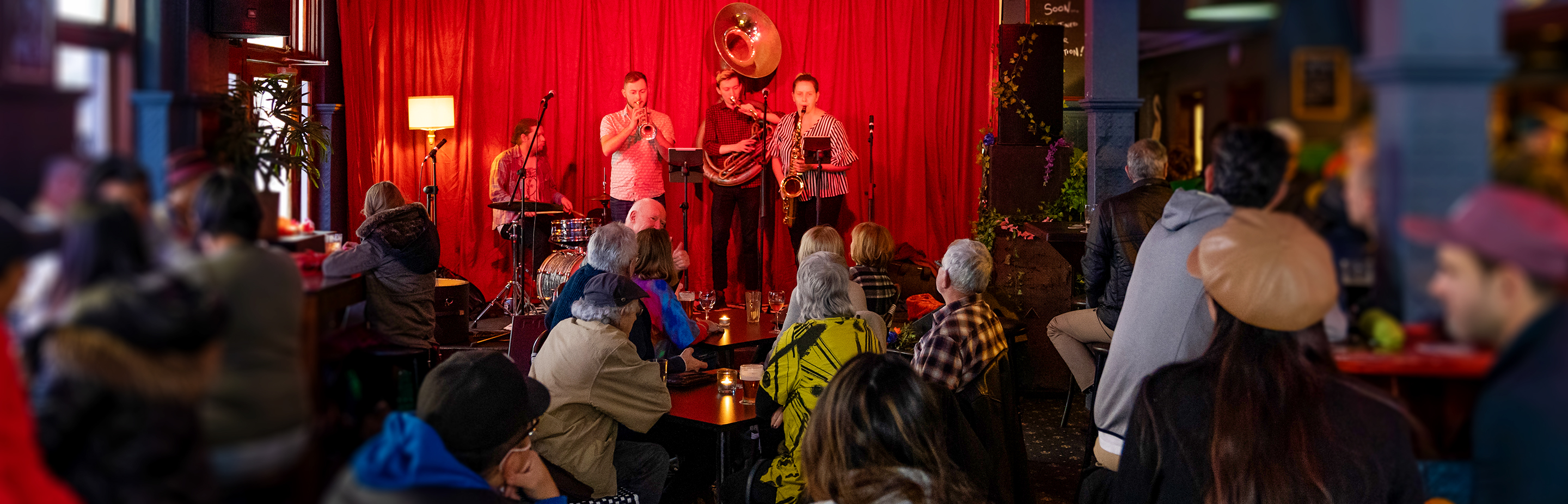 Band on a stage playing brass instruments in front of a red velvet curtain while an audience watches