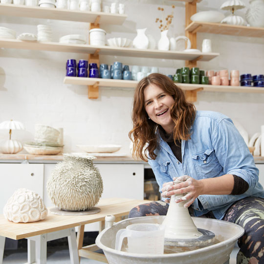 A smiling woman sits in front of a throwing wheel while moulding clay.