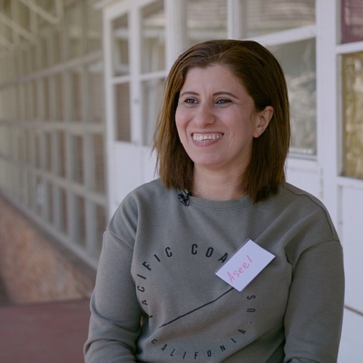 Photo of Aseel by Samuel Phibbs, Aseel sits in the Welcome Centre Courtyard, she wears an olive coloured jumper, has a big smile and a blond brown shoulder length haircut