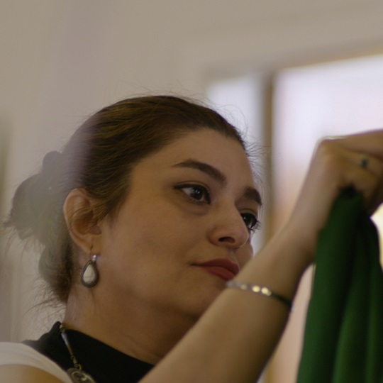 Photo of Elham by Samuel Phibbs, Elham looks at and holds a piece of green satin material, she wears a black and hite tshirt and her hair is pulled back in a low large bun, she has a tear drop pearl and silver coloured earring and a silver bracelet.