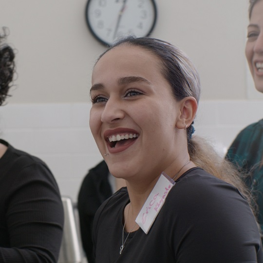 Photo of Erica by Samuel Phibbs, Erica smiles at the camera laughing her mouth is wide, another woman laughs in background. Erica wears a black top and has long blond hair with roots tied back in a wavy ponytail, she has a name tag fastened to her top with pink writing her name says Erica, she also has a face piercing above her lip