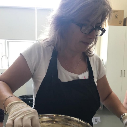 Photo of Ghada in the kitchen she wears a white tshirt with a black apron over the top, she has blong shoulder length hair and wears glasses.