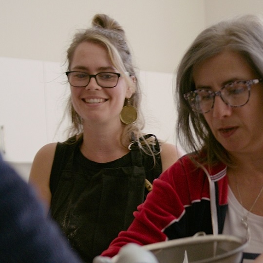 Photo of Raffaela in background she wears hair half up half down and has black frame rectangle glasses, she smiles and wears a black apron, to her left is Ghada another participant