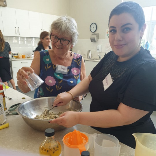 Photo of Suzie wearing a flowery top in a kitchen she is throwing ingredients in a large silver bowl, beside her is another project participant Maryam