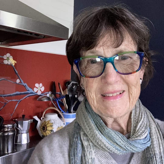 A photo of Tim, Tim has short grey hair and is in her kitchen in the background there is a stove top with red back splash and a coffee espresso machine