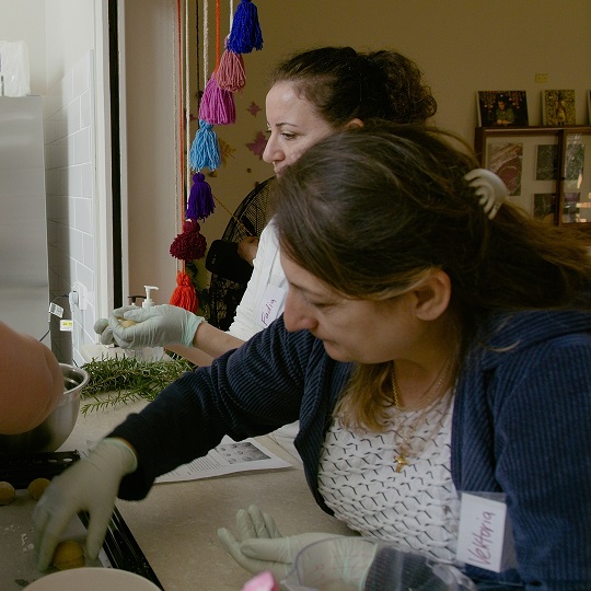 Photo of Vektoria in the kitchen she wears plastic serving gloves and places food on a plate, she has brown hair half up half down, she looks at the plate of food and workstation area