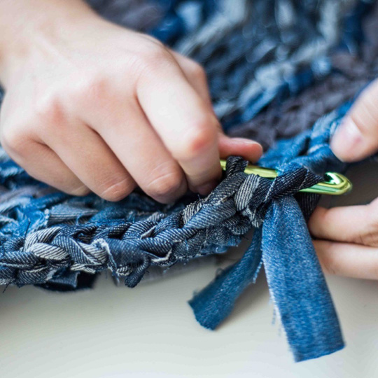 A close-up view of a person's hands knitting a weave of thick denim strips.