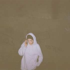 A woman in a hoodie stands against a brown wall