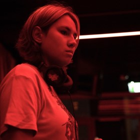 Side shot of a DJ wearing a light shirt, headphones around their neck and a short bob haircut standing under red lights
