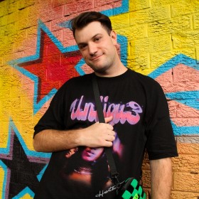 Young person standing against a colourful yellow wall with black and red stars wearing a black tshirt with pink and purple writing holding a satchel bag across the front of their body