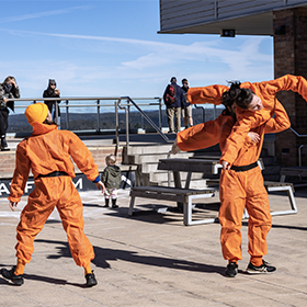 Three dancers in orange jumpsuits outside. One is being listed by another over their shoulder