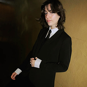 A woman in a black suit leaning against a brown wall