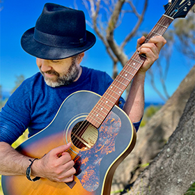 A man in a blue shirt and hat plays the guitar outside