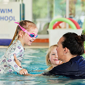 A woman in dark blue in the pool looks at a young child with goggles