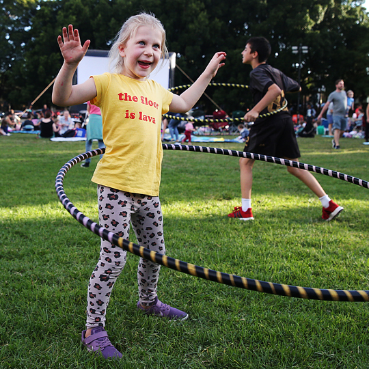 Young person with a yellow shirt and leopard print pants hula hooping on the grass with a big grin on their face