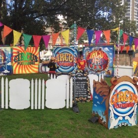 A colourful circus set up featuring rainbow bunting overhead, white picket fences and people practicing their juggling skills