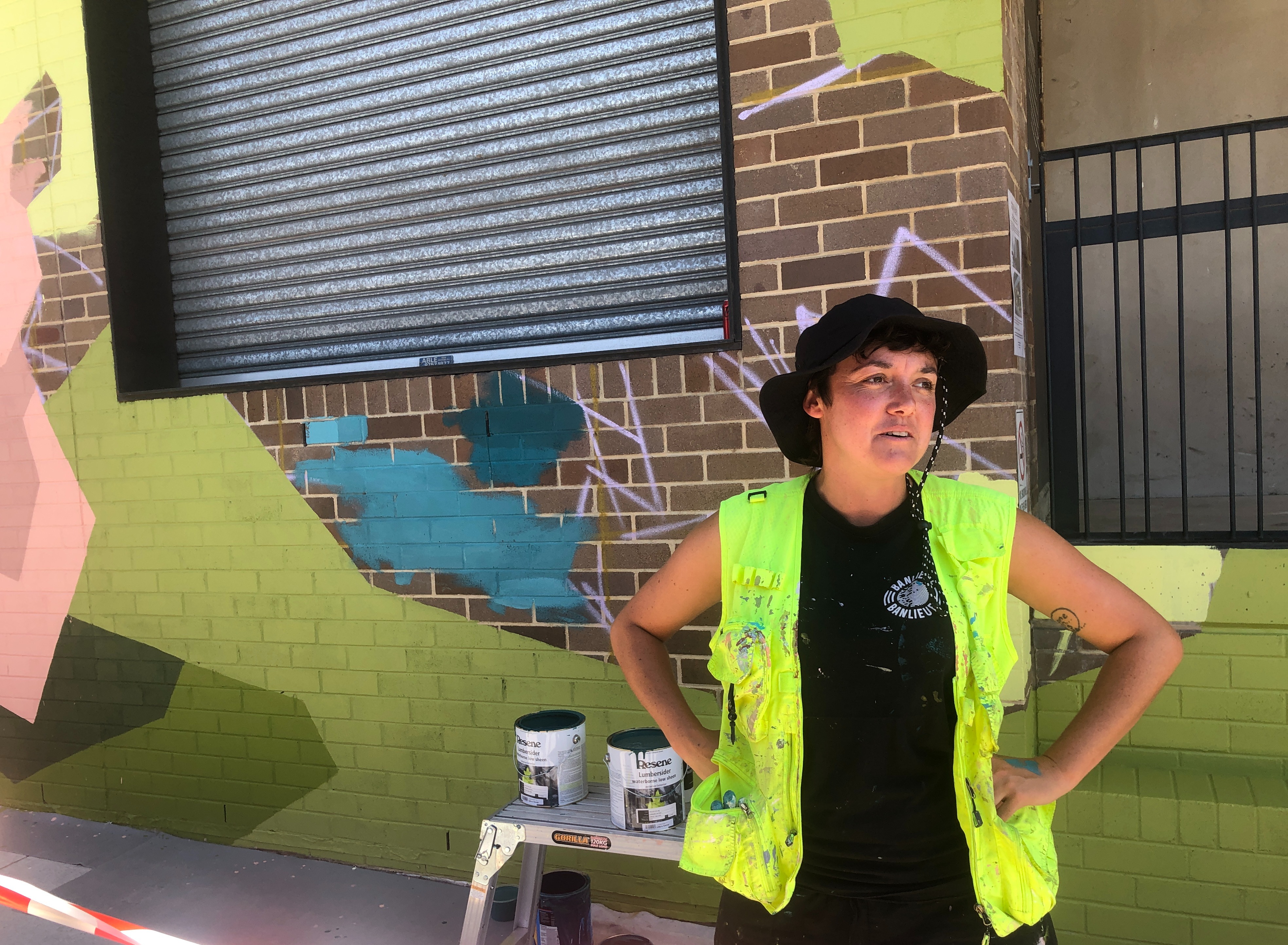 Woman in a high vis vest covered in multi-coloured paid stand in from out a large mural of geometric shaped painted in green, earthy tones