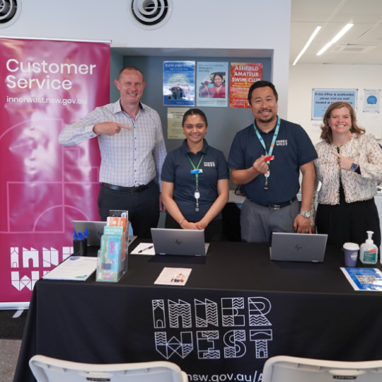 Four smiling pepople standing behid a trestle table next to a maroon pull-up banner that says Customer Service Inner West