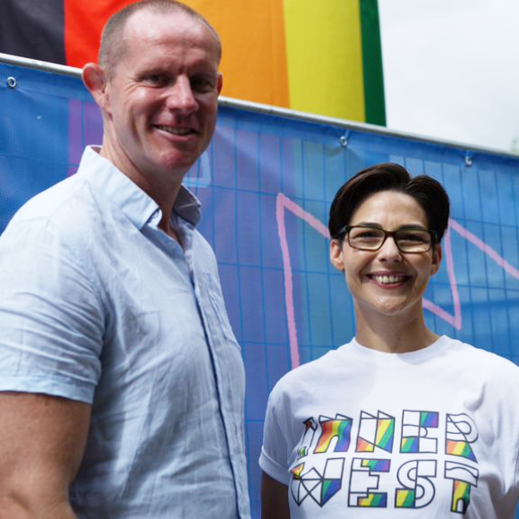 A man and a woman stand smiling in front of a struct wrapped in rainbow cladding. The woman's t-shirt reads 'Inner West' in rainbow colours
