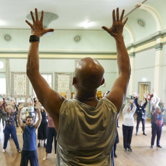 Seniors at a dance class- a bald man in the foreground raises his hands into the air, the crowd facing in copies his movement