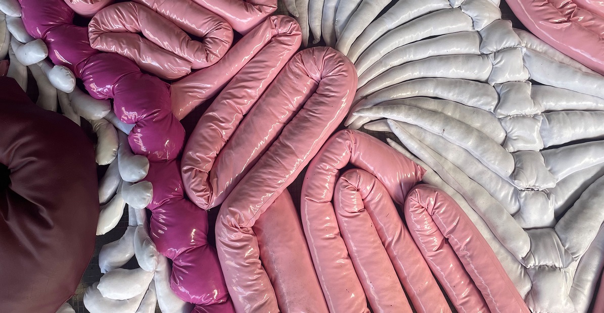 A contemporary textile art piece composed of glossy, padded fabric tubes arranged in intricate, overlapping patterns. The color palette includes pastel pink, fuchsia, white, and deep burgundy tones, resembling abstract, organic forms. The tubes are tightly coiled and layered, creating a plush, three-dimensional texture.