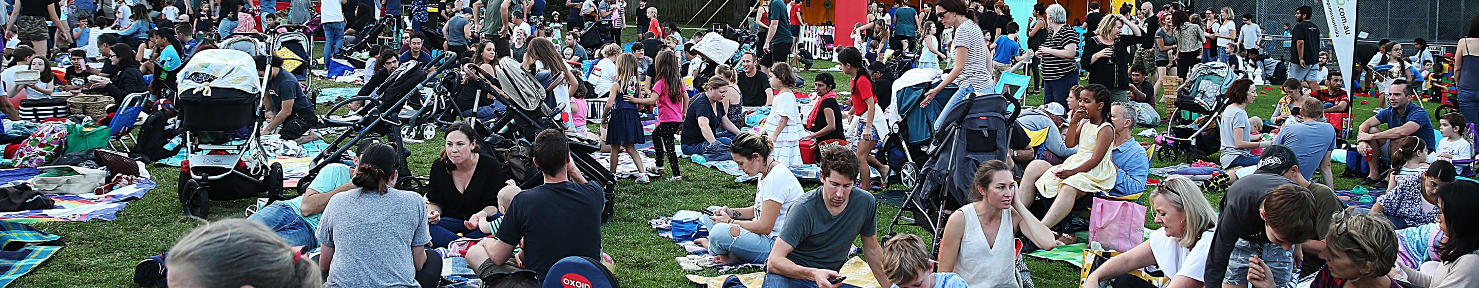 Large crowd of diverse people sitting in a park on picnic blankets facing to the left of the frame