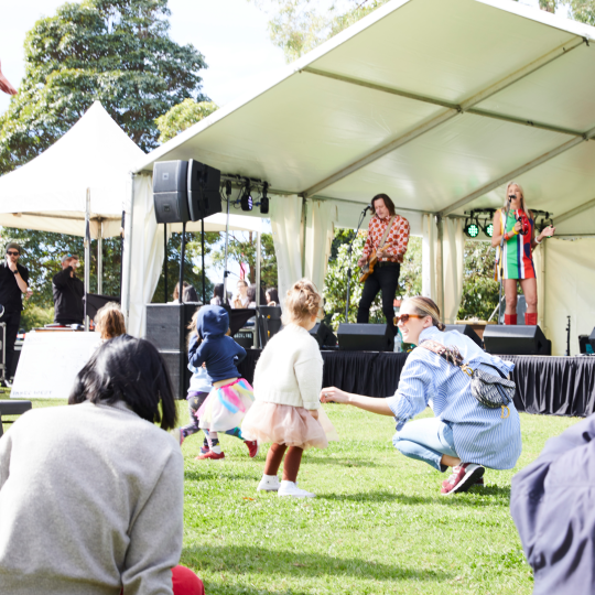 Park setting with a large stage featuring musicians and people young and old enjoying the entertainment from the grass in front