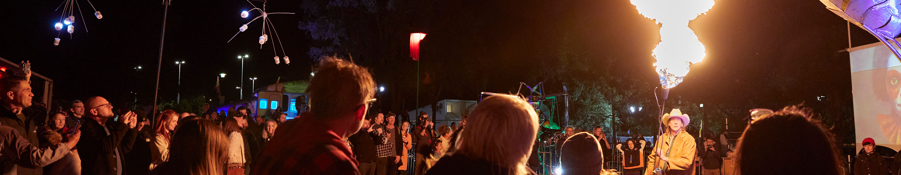 A crowd of people gathered at night under sculptural artworks, watching a man in a cowboy hat using a theatrical flamethrower