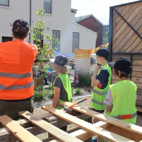 Group of people, young and old, wearing high vis jackets facing away from the camera surrounded by timber hand built items