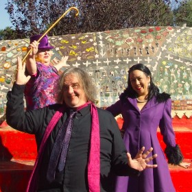 A group of three people brightly dressed in purple and pink standing in front of the oversized couch in Sydenham Green