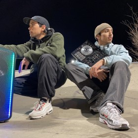 Two people sitting down in a skate park holding a large blue and green speaker and DJ deck