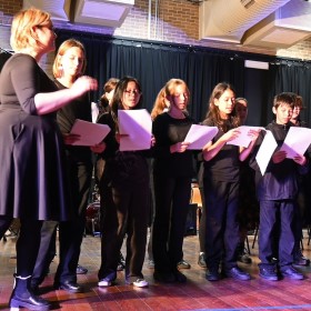 A student choir group standing and singing with sheet music along with their conductor