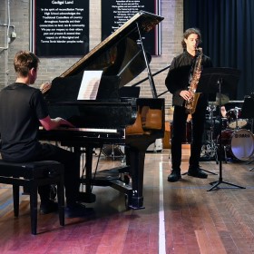 A school hall setting featuring someone playing a grand piano and saxophone with a drum kit in the background