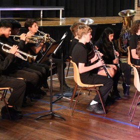 A group of students in a school hall setting sitting down playing a variety of wind instruments