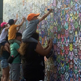 A group of three adults all with children on their shoulders drawing onto a wall covered in cat faces