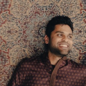 A young man wearing a patterned maroon collared shirt standing against a decorative backdrop looking to the right