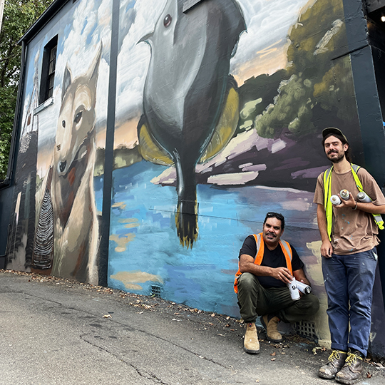 two smiling artists stand in front of a mural on the side of a house.